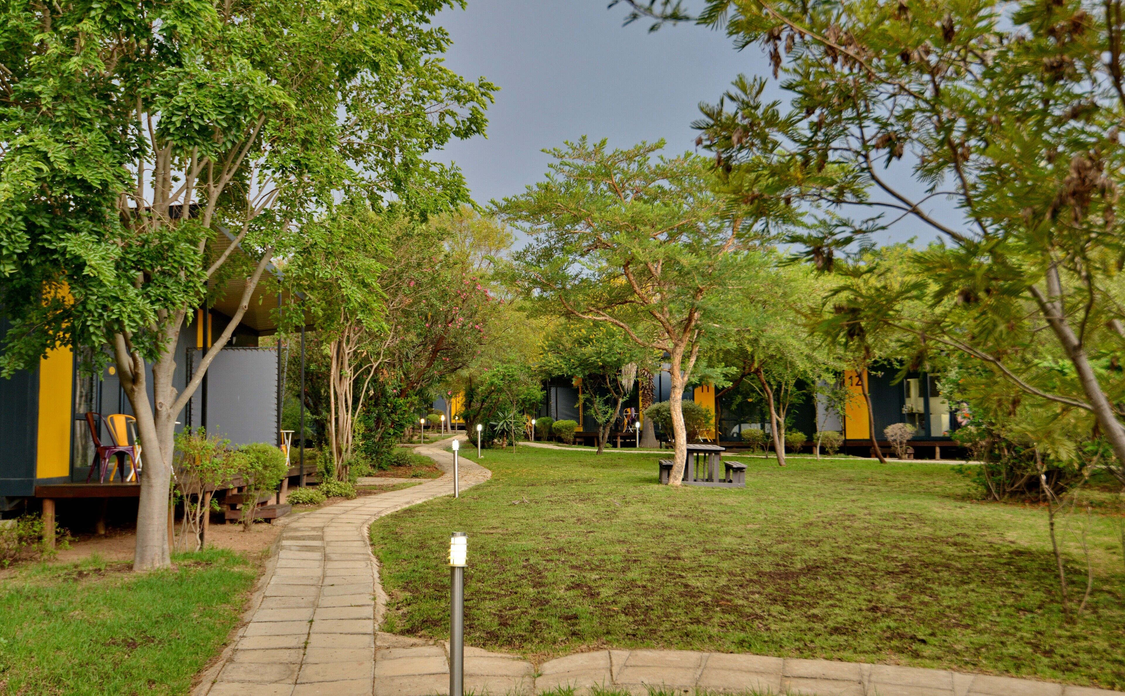 A pathway leading through a garden area with trees and a building with yellow and gray walls.
