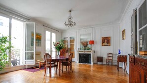 Une pièce spacieuse dotée d’un parquet en bois disposé en chevrons, d’une table à manger avec des chaises, d’un lustre, d’une cheminée et d’une grande fenêtre offrant une vue dégagée.