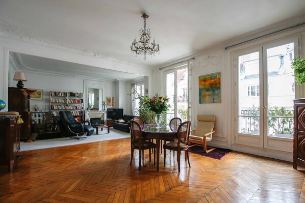 Ein geräumiges Wohnzimmer mit Holzparkett, einem Kronleuchter, einem Esstisch mit Stühlen, einem Bücherregal und einem Fernseher.