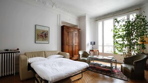 Une pièce bien éclairée, avec un lit, un canapé, une chaise, une armoire en bois et une grande fenêtre agrémentée d’une plante.
