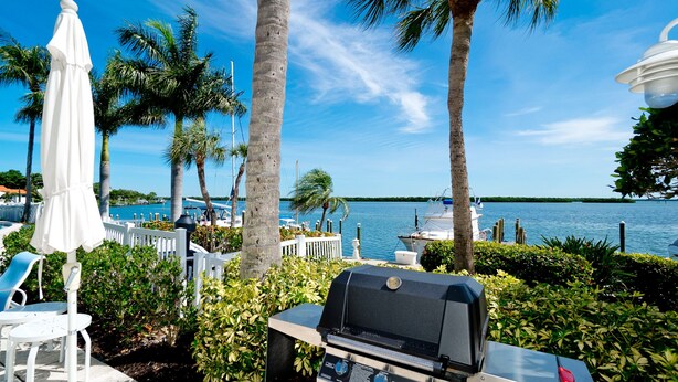A patio with a grill, white chairs, and umbrellas overlooking a body of water with boats. - GORGEOUS UPSCALE LONGBOAT KEY CONDO OFFERS BAYFRONT VIEWS!! (Longboat Key)