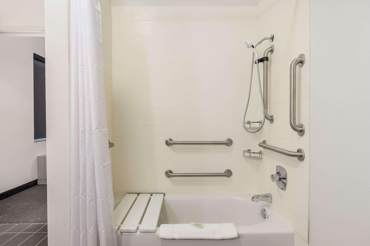 A bathroom with a white bathtub, grab bars, a shower head, and a towel on the floor.