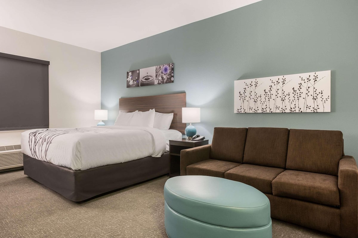 A hotel room with a bed, a brown sofa, a blue ottoman, and a nightstand with a lamp.