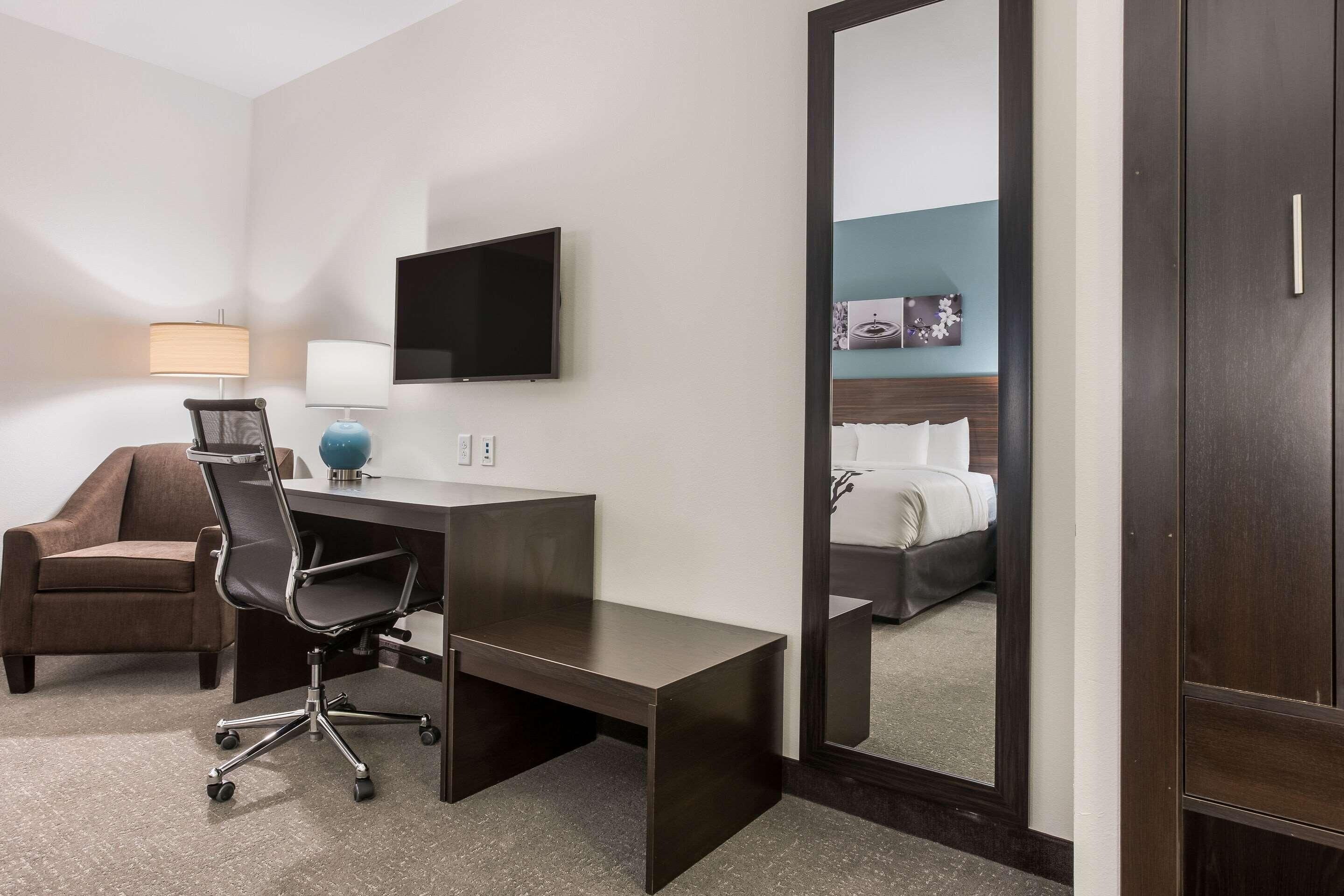 A hotel room with a desk, chair, TV, and a large mirror reflecting a bed.