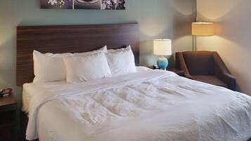 A neatly made bed with white linens, a wooden headboard, two bedside lamps, a chair, and a wall-mounted artwork.