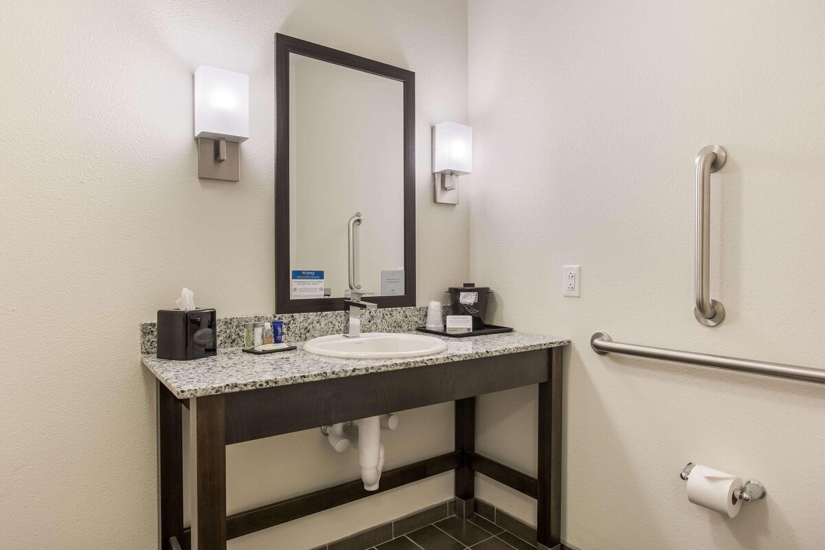 A bathroom with a sink, mirror, towel dispenser, and handrail.