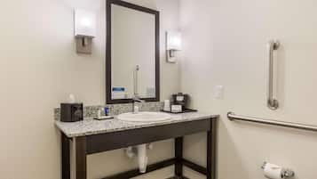 A bathroom with a sink, mirror, towel dispenser, and handrail.