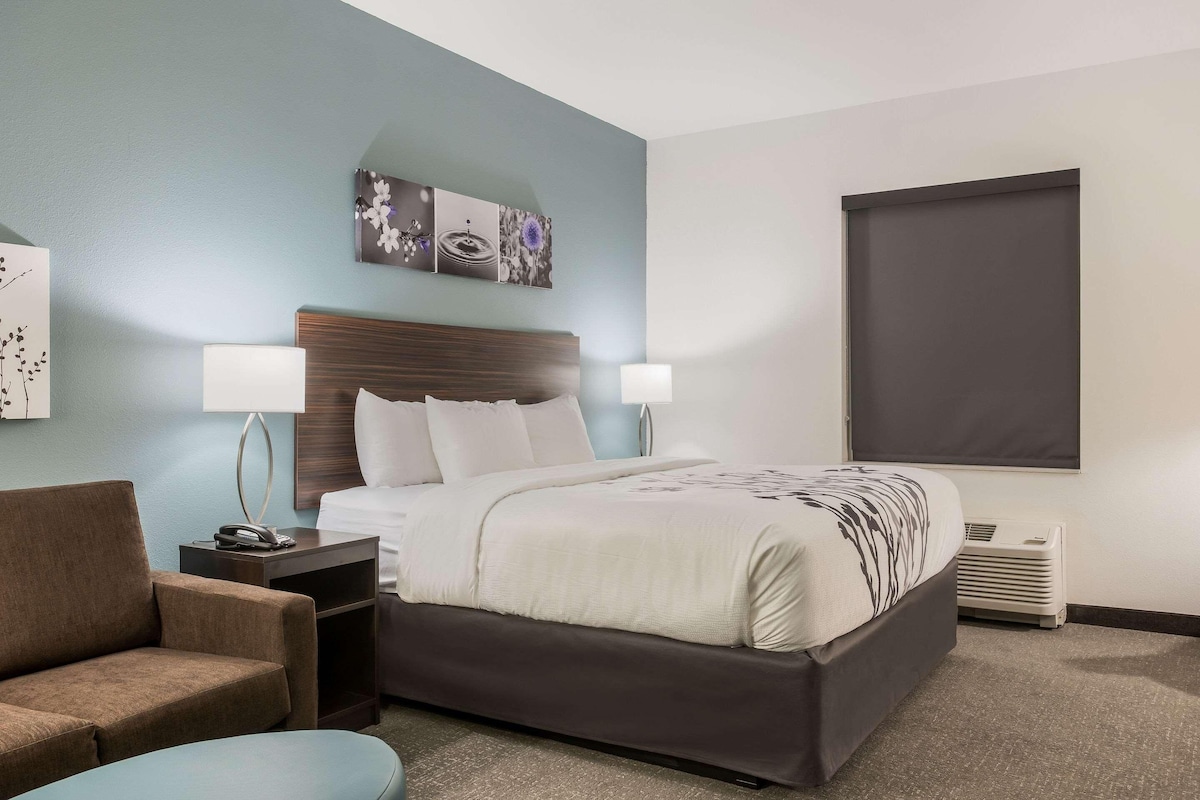 A hotel room with a bed, two bedside lamps, a nightstand, a chair, and a wall-mounted artwork.