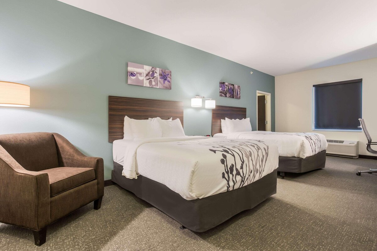 A hotel room with two beds, a brown armchair, a desk, and a window.