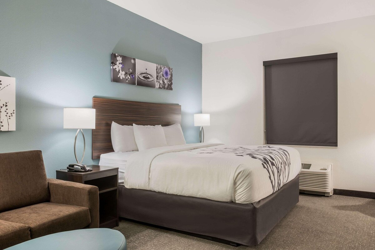 A hotel room with a bed, two bedside lamps, a nightstand, a chair, and a wall-mounted artwork.