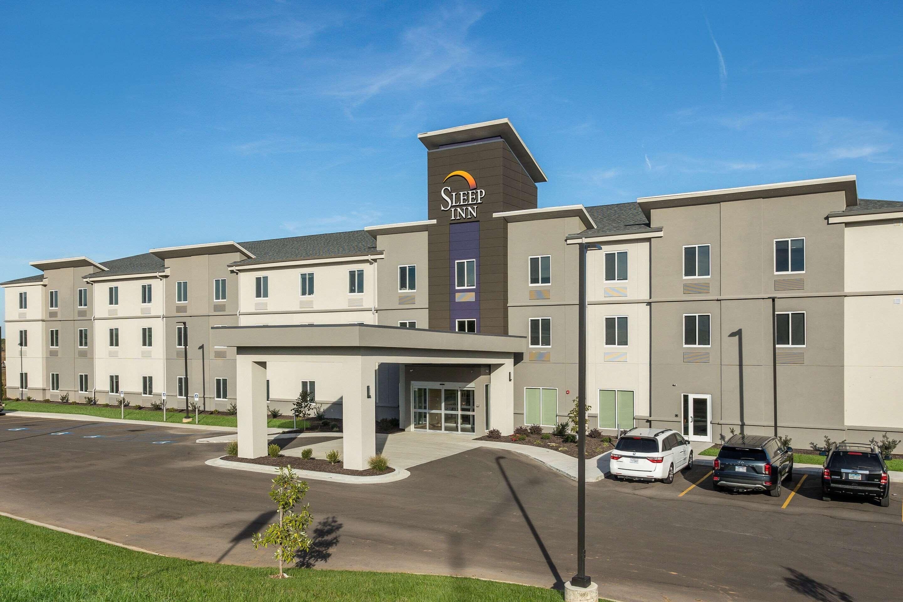 A Sleep Inn hotel with a modern design, featuring a central entrance, multiple parking spaces, and a clear sky.