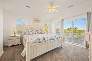 A bedroom with a bed, a nightstand, a lamp, a ceiling fan, and a large window with a view of the outdoors.