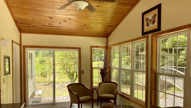 A spacious living room with a wooden ceiling, large windows, a ceiling fan, and a view of a garden.