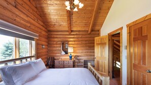 Un dormitorio tipo cabaña de madera con techo de madera, una cama con sábanas blancas, un aparador, un espejo y una puerta que da a otra habitación.