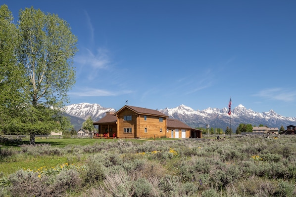 Une cabane en bois dotée d’un porche, entourée d’un champ verdoyant et avec des montagnes en arrière-plan.