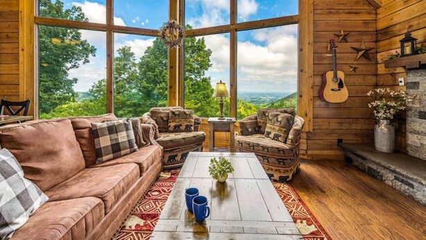 Interior acogedor de una cabaña con un ventanal que ofrece vistas panorámicas, una chimenea y muebles rústicos.