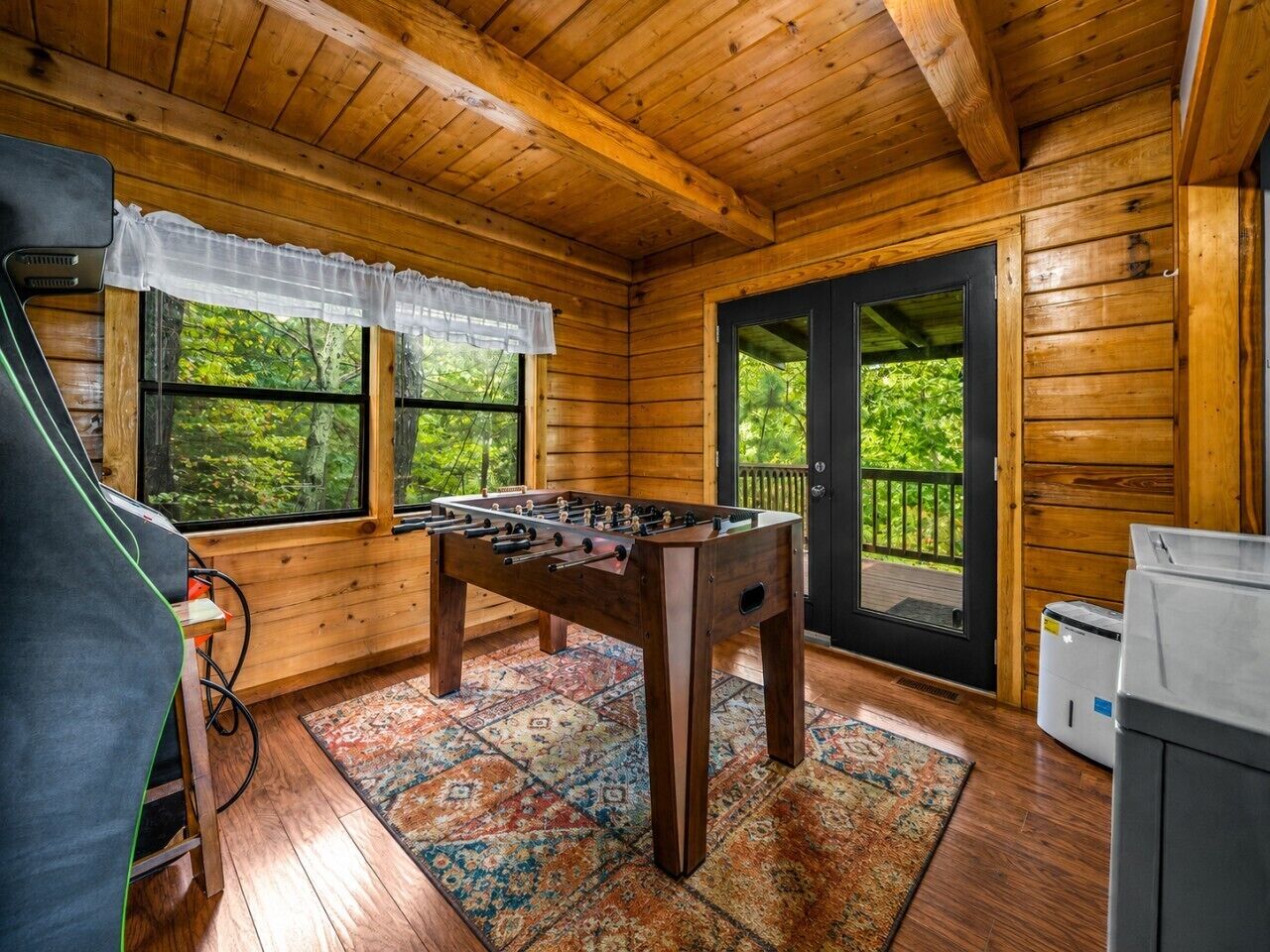 A wooden-floored room with a foosball table, a window with a view of greenery, and a door leading outside.