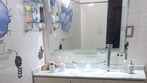 Une salle de bain avec un lavabo en verre, un miroir et des toilettes. On y trouve des étagères avec des articles de toilette et des serviettes.