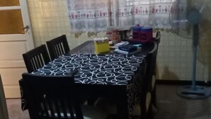 Un coin repas avec une table dressée pour quatre personnes, des chaises, un ventilateur et une fenêtre agrémentée de rideaux à motifs.
