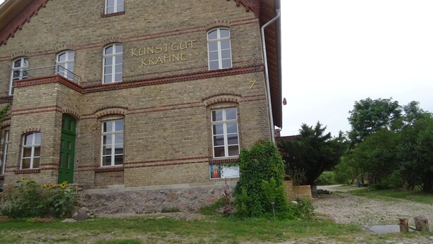 A historic building with the name "Kunstgut Krahn" on the facade, featuring a stone exterior, multiple windows, and a small garden area with sunflowers. - Vacation apartment "Biene" in the listed Krahne art estate (Kloster Lehnin OT  Krahne)