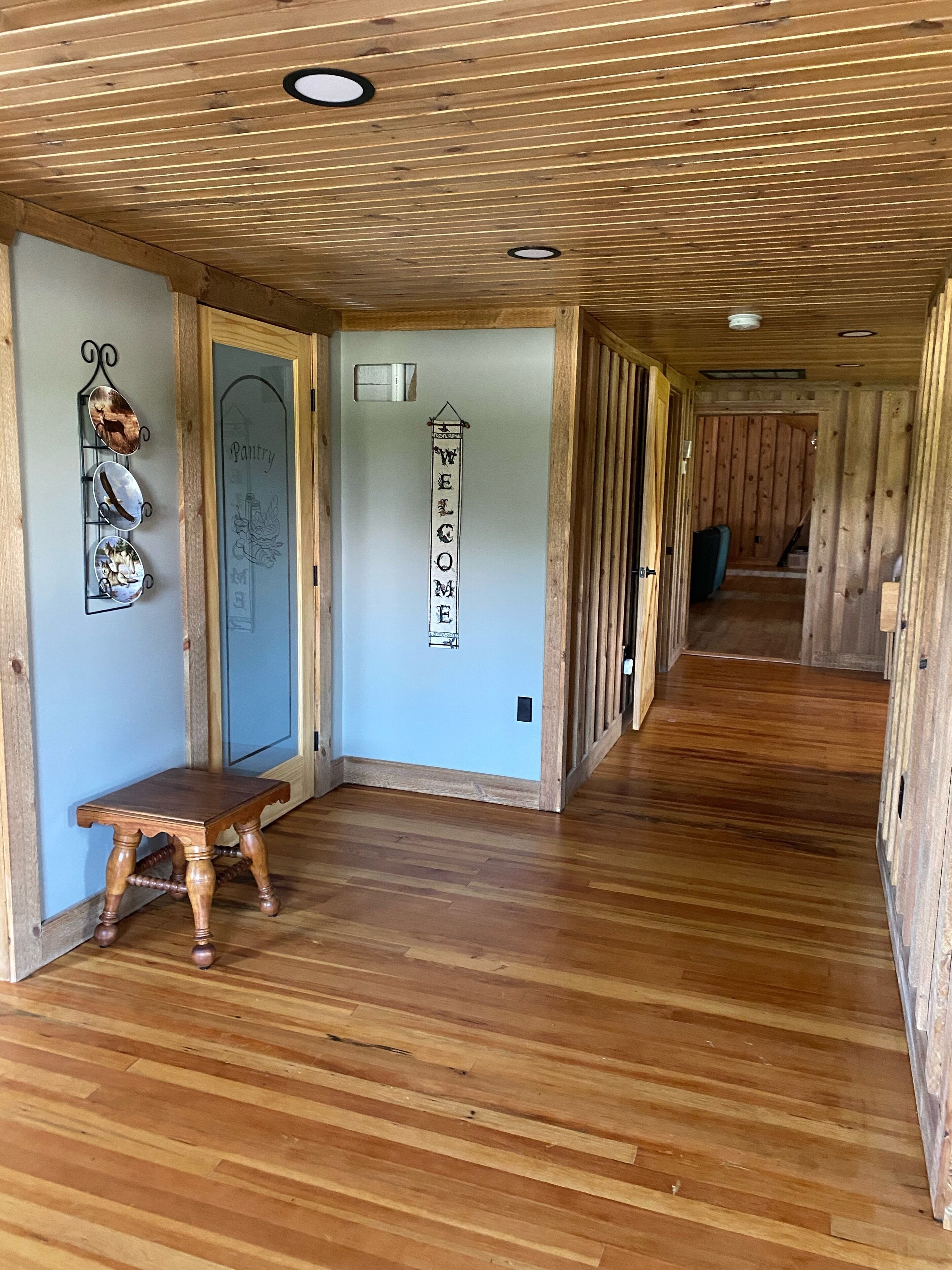 A wooden interior hallway with a 'Welcome' sign, a small table, and a door labeled 'Pantry'.