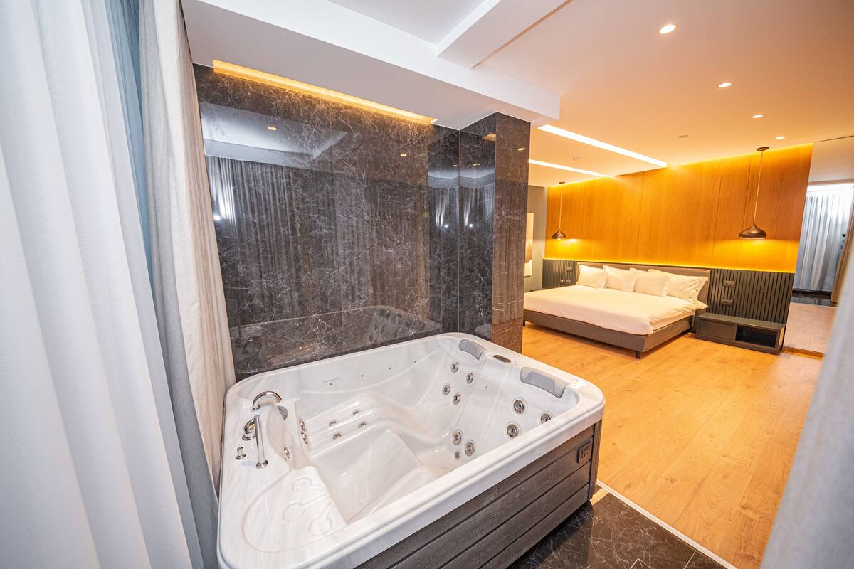 Une salle de bain moderne avec une grande baignoire, un plancher en bois et un lit avec une tête de lit en bois dans la pièce adjacente.