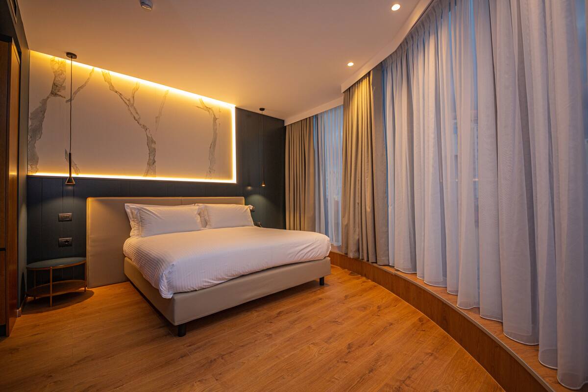 Une chambre moderne avec un grand lit, un plancher en bois, un luminaire fixé au mur et des rideaux légers.
