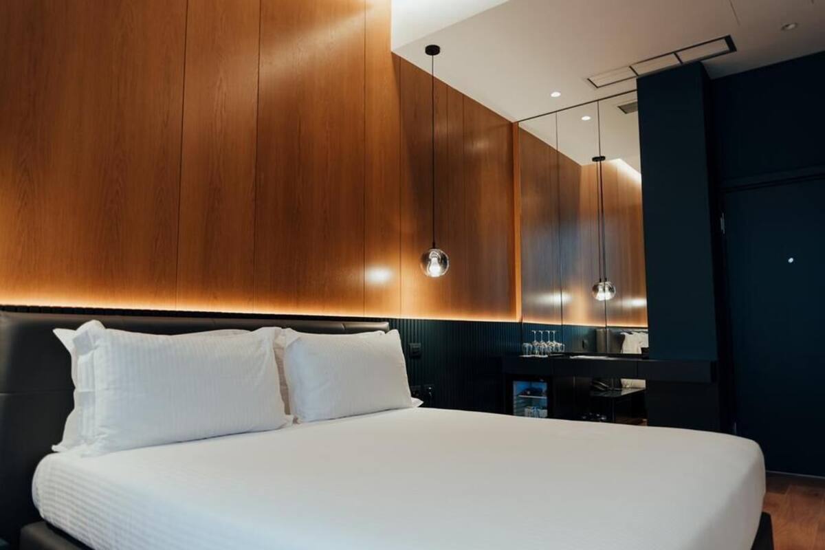 Une chambre d’hôtel moderne avec un grand lit, des murs recouverts de panneaux en bois, une tête de lit de couleur sombre et un petit coin cuisine.