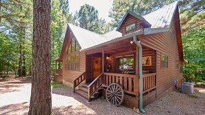 A rustic cabin with a porch, a wooden wheel, and a gravel driveway surrounded by trees. - TEXAS 2 STEP Luxury Log Cabin at the Entrance to Beavers Bend State park (Broken Bow)