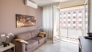 Un salón moderno con un sofá, un cuadro, una planta en maceta y vistas a un balcón exterior a través de un ventanal.