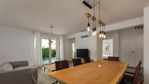 Ett modernt vardagsrum med ett stort matbord i trä, stolar i rotting, en grå soffa och en tv.