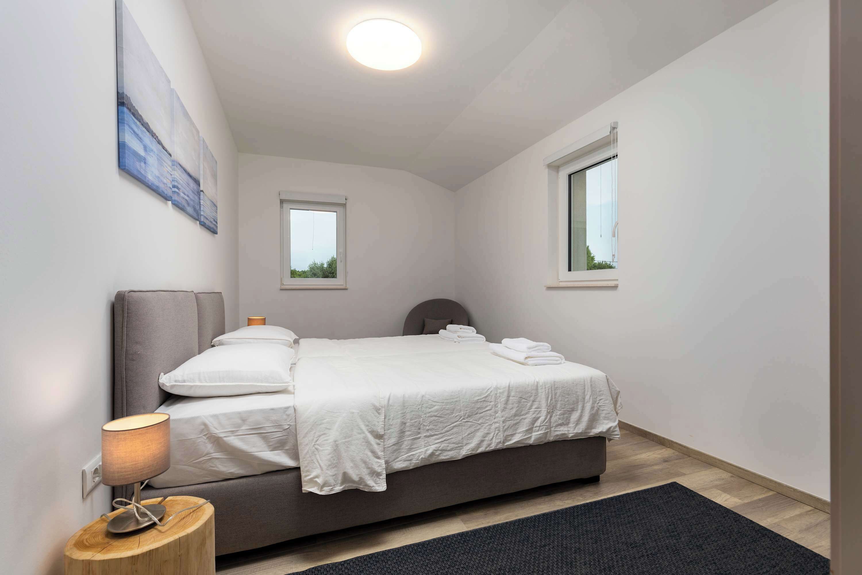 A modern bedroom with a bed, bedside lamp, and two windows.