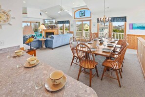 Un vaste espace de vie comprenant un coin repas avec une table dressée pour six personnes, entourée de chaises en bois. Une grande fenêtre offre une vue sur le quartier.