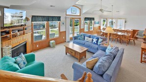 Uma sala de estar espaçosa com um sofá azul grande, uma mesa de centro de madeira e uma televisão sobre um suporte de madeira. Há um espaço de jantar com cadeiras e mesa de madeira, e uma varanda com grades de madeira.