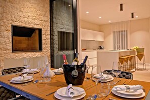 Una mesa de comedor preparada con platos, cubiertos y botellas de vino en una habitación moderna con pared de piedra y una cocina al fondo.