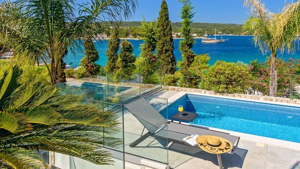 A glass-enclosed infinity pool with a lounge chair and table, overlooking a coastal area with trees and a distant boat.