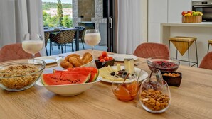 A breakfast table with various food items and drinks, a view of the outdoors, and a modern kitchen in the background.