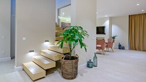 A modern living room with a staircase, a large potted plant, a flat-screen TV, and a dining area.