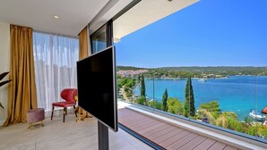 A balcony with a TV, a red chair, a small ottoman, and a view of a coastal town and sea.