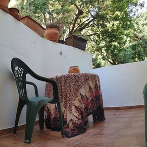 A small outdoor patio with a green plastic chair, a table covered with a patterned tablecloth, and terracotta pots on a white wall.