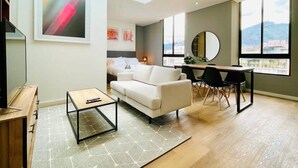 A modern living room with a white sofa, a coffee table, and a dining area with chairs. There is a large window providing a view of the city. - w*| Modern Duplex in Parque Virrey (Bogotá)