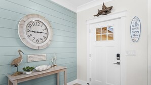 A hallway with a wooden console table, a wall clock, a decorative bird, a small plant, and a door with a window. - Spacious Beachfront 3 Bedroom! (Destin)