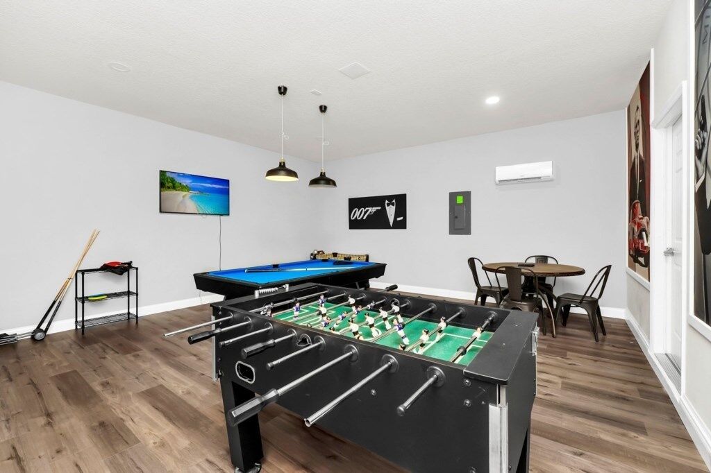 A room with a foosball table, a pool table, and a billiards table. There are two pendant lights, a painting of a beach, and a framed picture on the wall.
