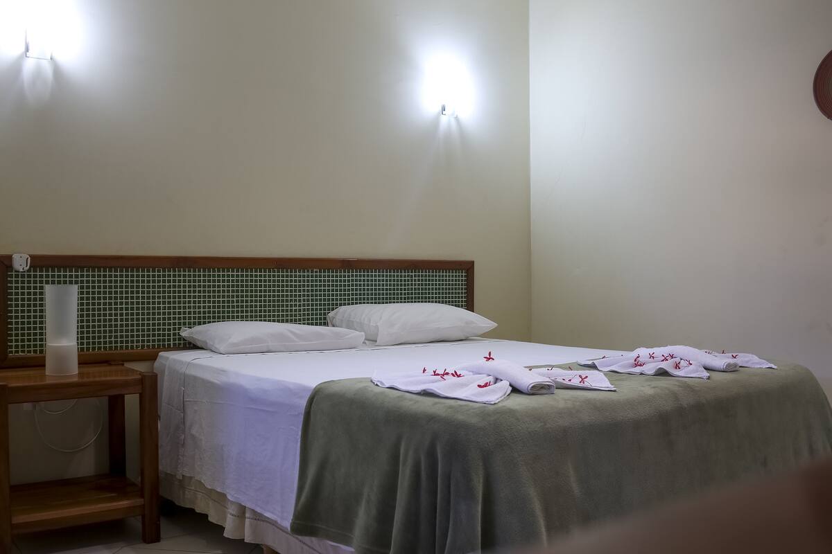 A neatly made bed with white linens and a green bedspread, a wooden nightstand with a lamp, and a wall-mounted light fixture.