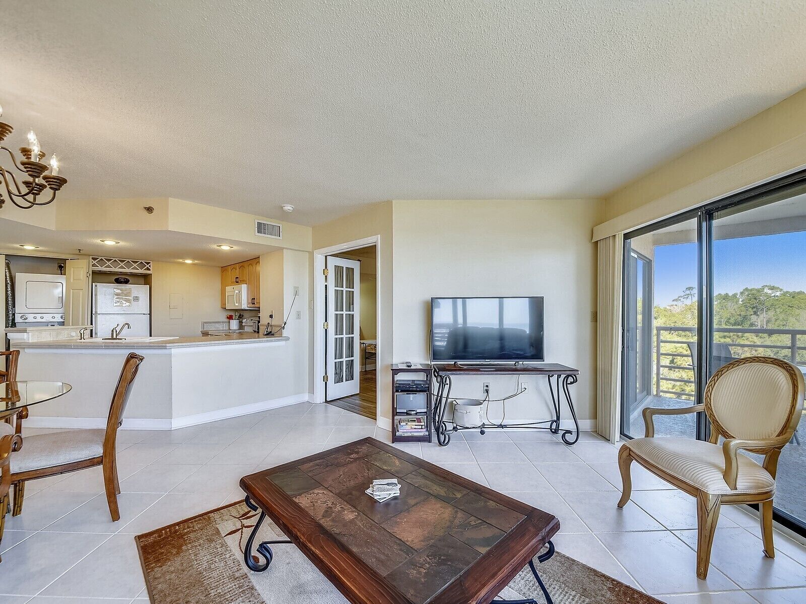 A spacious living area with a kitchen, a dining table, chairs, a coffee table, a TV, and a balcony with a view.