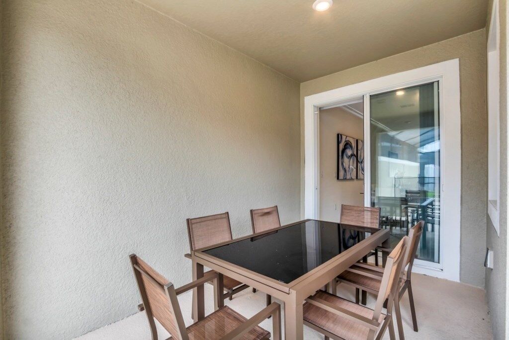 A dining area with a glass-top table and wooden chairs, a sliding glass door leading to an outdoor space, and a light-colored wall.