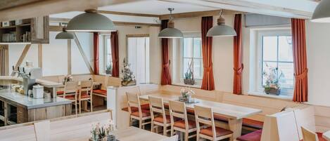 Uma ampla área de jantar com várias mesas e cadeiras, vigas de madeira no teto e grandes janelas com cortinas.