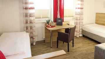 A room with a bed, a desk with a chair, a small table with a plant, and a window with curtains.