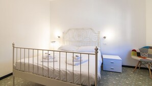 Un dormitorio con estructura de cama de metal blanco, una mesita de noche con cajones, una silla con cojín colorido y una lámpara de pared.