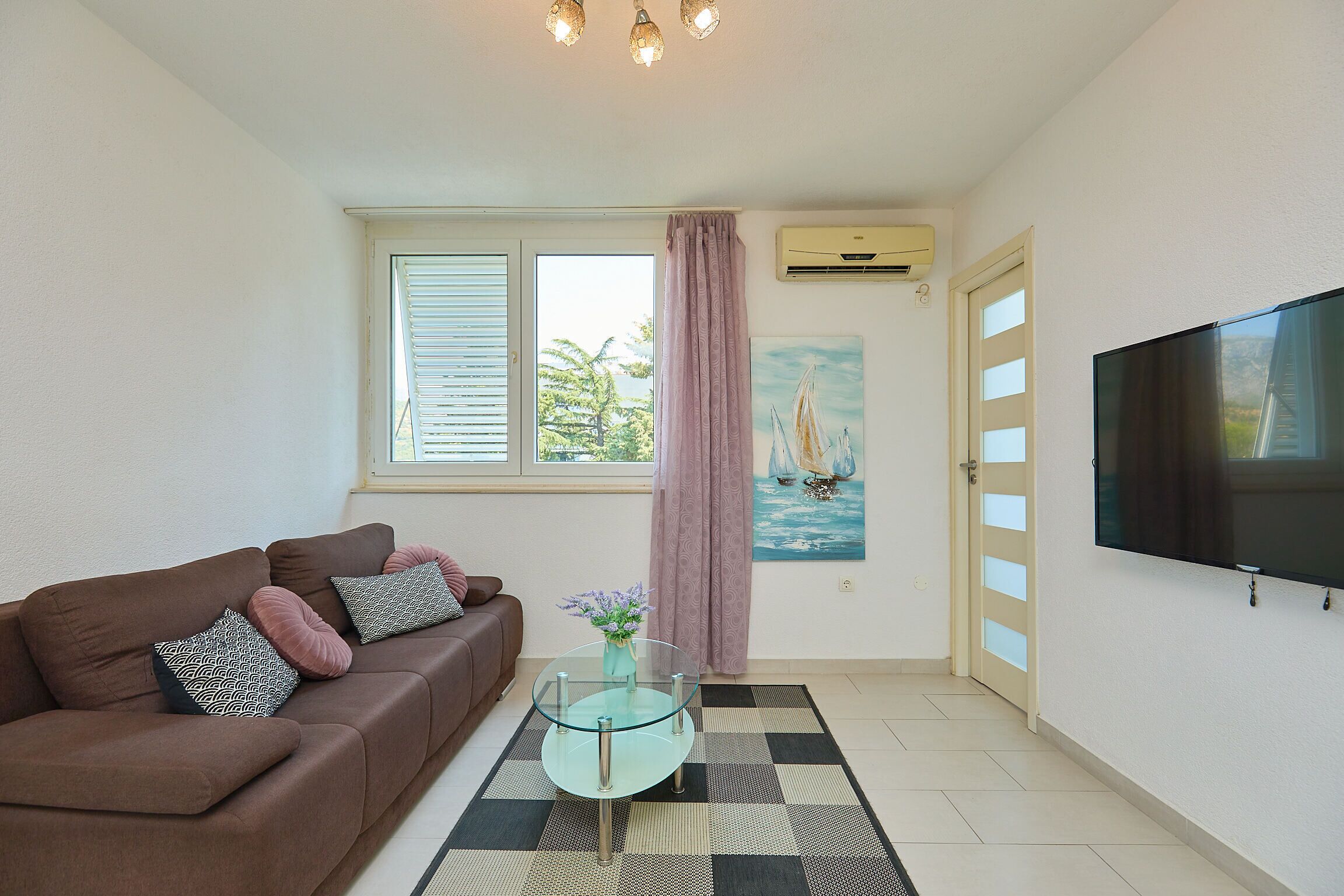 A living room with a brown sofa, a glass coffee table, a painting of a sailboat, and a TV on the wall.